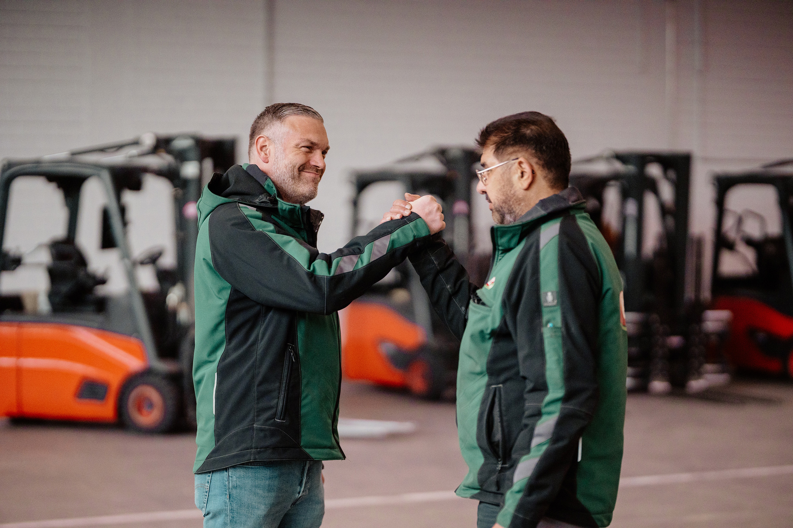 Two employees greeting each other in front of forklifts in a warehouse.